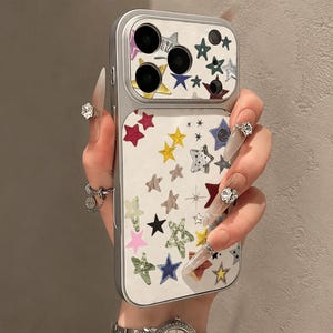 May include: A silver-toned smartphone case featuring a white back panel decorated with a scattered pattern of multi-colored stars in various textures and designs, including solid, patterned, and outlined shapes, surrounding the camera lens array.