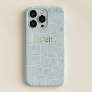 Peut inclure: Coque de téléphone bleu clair avec une texture imitation peau de crocodile. Le boîtier comporte trois objectifs d'appareil photo et le mot "Deb" en lettres dorées. Coque pour smartphone.