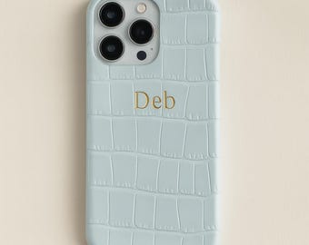 May include: A light blue phone case with a crocodile skin texture. The case has three camera lenses and the word "Deb" in gold lettering. The phone case is designed to fit a smartphone.