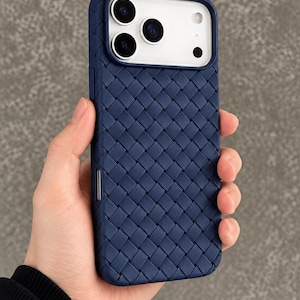 Könnte beinhalten: Eine marineblaue Smartphone-Hülle mit einer geflochtenen Korbgeflecht-Struktur auf der Rückseite. Die Hülle hat eine rechteckige Aussparung an der Oberseite für ein Dreifach-Kamerasystem und einen Blitz sowie einen schmalen Metallknopf an der Seite.