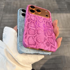 May include: Two smartphone cases featuring a textured snakeskin pattern. One case is bright pink with a metallic copper-toned camera frame, while the other is light blue with a silver-toned camera frame. Both cases have cutouts for the camera lenses.