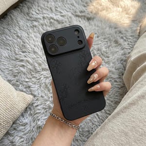 May include: Black phone case with a raised camera section and embossed cross designs. The case has the text "CHROME HEARTS" at the bottom. The phone case is held in a hand.