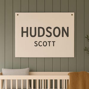 May include: A cream-colored sign with the name "HUDSON" above "SCOTT" in dark gray letters. The sign is mounted on a green wall above a wooden crib with a gray pillow and a mustard-colored blanket.