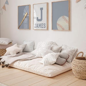 K&ouml;nnte beinhalten: Ein wei&szlig;-graues Bodenkissen mit einer grauen Decke und mehreren wei&szlig;en und grauen Kissen. An der Wand hinter dem Kissen h&auml;ngen drei gerahmte Drucke. Die Drucke zeigen einen Baseballschl&auml;ger, den Buchstaben J und den Namen James sowie einen Baseball.