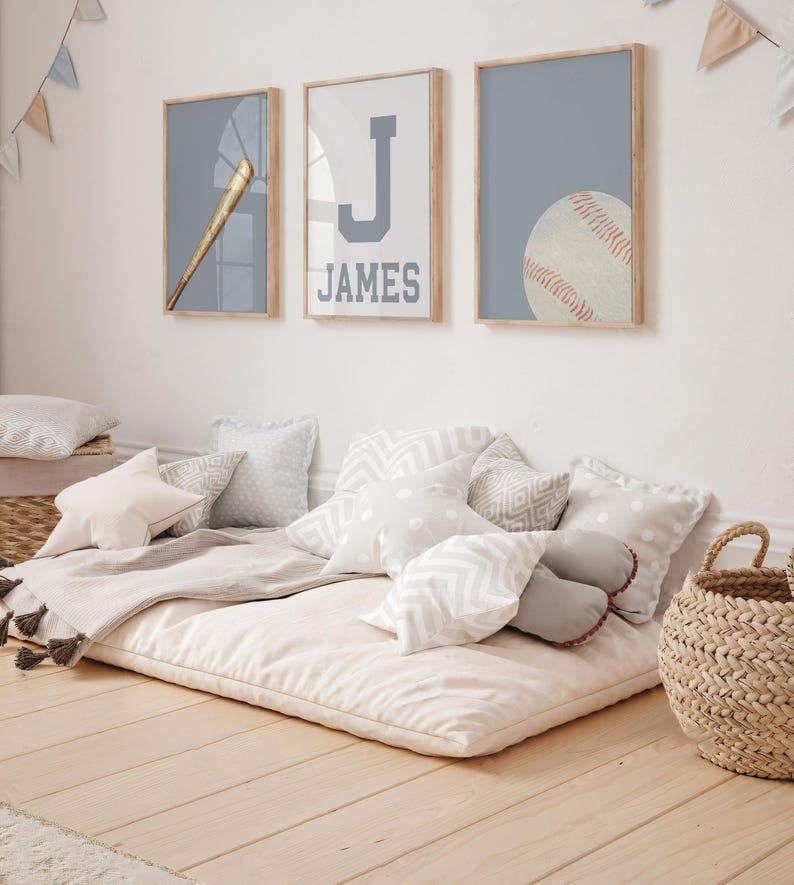 K&ouml;nnte beinhalten: Ein wei&szlig;-graues Bodenkissen mit einer grauen Decke und mehreren wei&szlig;en und grauen Kissen. An der Wand hinter dem Kissen h&auml;ngen drei gerahmte Drucke. Die Drucke zeigen einen Baseballschl&auml;ger, den Buchstaben J und den Namen James sowie einen Baseball.