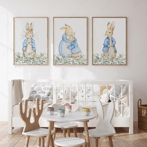 May include: Three framed watercolor prints featuring Peter Rabbit characters. Each print shows a rabbit in a blue jacket, set against a backdrop of green foliage. The prints are displayed above a white crib.