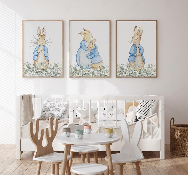 May include: Three framed watercolor prints featuring Peter Rabbit characters. Each print shows a rabbit in a blue jacket, set against a backdrop of green foliage. The prints are displayed above a white crib.