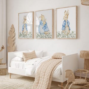May include: Three framed watercolor prints featuring Peter Rabbit in a blue jacket, set against a white background with green foliage. The prints are displayed above a white bed with beige bedding, creating a nursery theme.