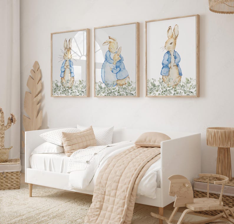 May include: Three framed watercolor prints featuring Peter Rabbit in a blue jacket, set against a white background with green foliage. The prints are displayed above a white bed with beige bedding, creating a nursery theme.