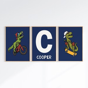 May include: Three framed prints with a blue background. The center print features a white letter C and the word "COOPER" below. The left print shows a green dinosaur riding a red bicycle. The right print shows a green dinosaur riding a skateboard.