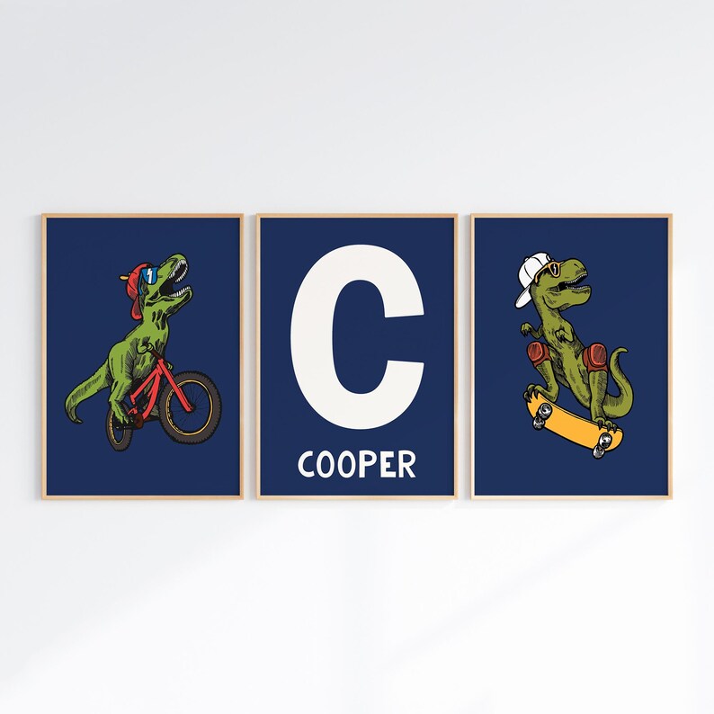 May include: Three framed prints with a blue background. The center print features a white letter C and the word "COOPER" below. The left print shows a green dinosaur riding a red bicycle. The right print shows a green dinosaur riding a skateboard.