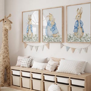 May include: Three framed watercolor prints of Peter Rabbit in a nursery setting. Each print features a different scene of Peter Rabbit in a blue jacket. A woven giraffe and storage unit with pillows and bins complete the room's decor.