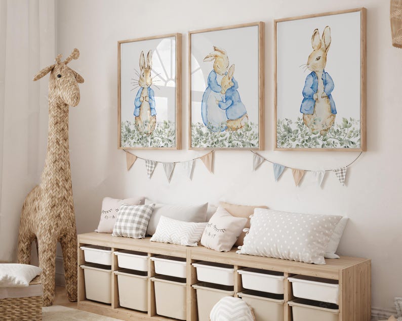 May include: Three framed watercolor prints of Peter Rabbit in a nursery setting. Each print features a different scene of Peter Rabbit in a blue jacket. A woven giraffe and storage unit with pillows and bins complete the room's decor.