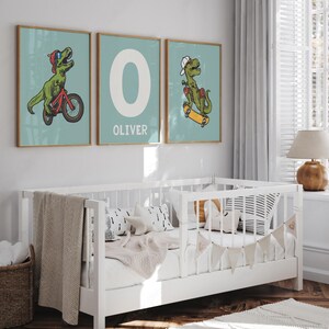May include: Three framed prints with a light blue background. The prints feature a green dinosaur riding a bicycle, the letter O, and a green dinosaur riding a skateboard. The letter O print has the name Oliver written below it.