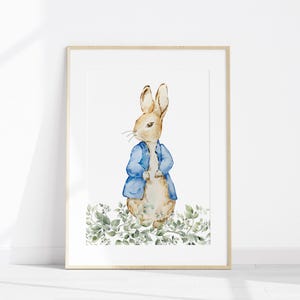 May include: Watercolor print of a rabbit wearing a blue jacket, standing amidst green foliage. The artwork is framed in a light wood frame, perfect for nursery or children's room decor. The rabbit is the central focus of the artwork.