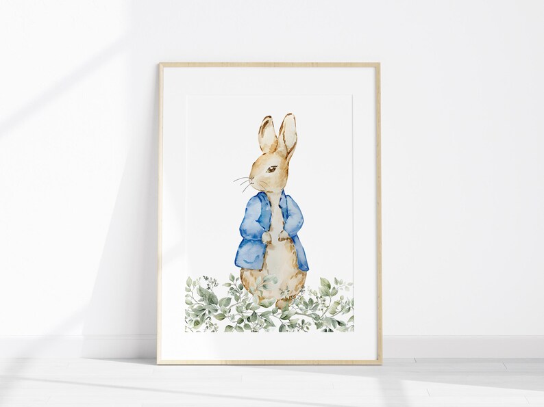 May include: Watercolor print of a rabbit wearing a blue jacket, standing amidst green foliage. The artwork is framed in a light wood frame, perfect for nursery or children's room decor. The rabbit is the central focus of the artwork.