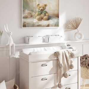 May include: A nursery scene featuring a framed painting of a teddy bear with a green bow tie. A white dresser with a changing pad, a pom-pom blanket, and a woven basket are in the foreground. A white bunny figurine and a vase with a dried palm frond are also visible.