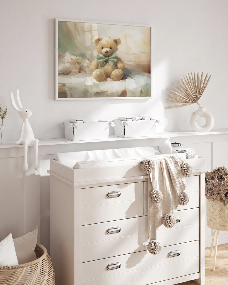 May include: A nursery scene featuring a framed painting of a teddy bear with a green bow tie. A white dresser with a changing pad, a pom-pom blanket, and a woven basket are in the foreground. A white bunny figurine and a vase with a dried palm frond are also visible.