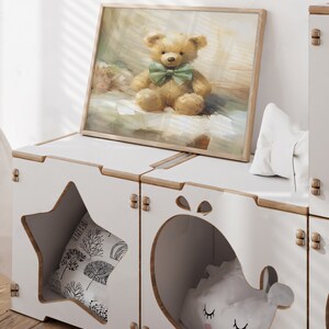 May include: A framed print of a teddy bear with a green bow tie sits atop a white storage unit. The unit has star and whale-shaped cutouts, revealing pillows inside. The print and unit create a cozy, whimsical aesthetic.