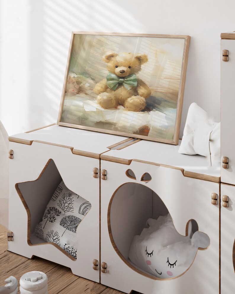 May include: A framed print of a teddy bear with a green bow tie sits atop a white storage unit. The unit has star and whale-shaped cutouts, revealing pillows inside. The print and unit create a cozy, whimsical aesthetic.