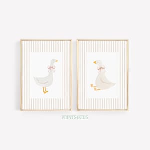 May include: Two framed prints featuring watercolor illustrations of white geese with pink bows around their necks. The geese are set against a striped background, with gold frames. The text "PRINTS4KIDS" is at the bottom.