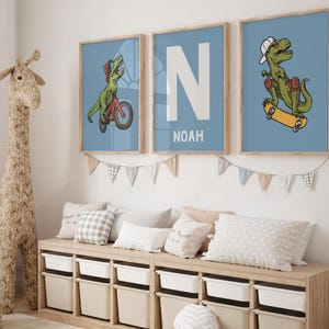 May include: Three framed prints with a blue background. The prints feature a dinosaur riding a skateboard, a dinosaur riding a bicycle, and the letter N with the name Noah underneath.