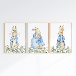 May include: Three framed watercolor prints featuring rabbits in blue jackets and dresses. The rabbits are set against a backdrop of green foliage. The artwork has a soft, whimsical style, perfect for a nursery or children's room.