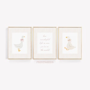 May include: A set of three framed prints with gold frames, each displaying a watercolour illustration of a goose with a pink bow. The prints have a striped background. The middle print has the text "how wonderful life is now you're in the world".