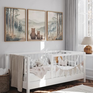 May include: A white cot with a beige blanket and decorative pillows is positioned below three framed watercolour paintings of a forest scene. The paintings depict a family of bears. A wicker basket is on the floor.