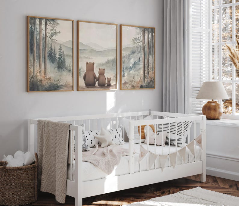 May include: A white cot with a beige blanket and decorative pillows is positioned below three framed watercolour paintings of a forest scene. The paintings depict a family of bears. A wicker basket is on the floor.