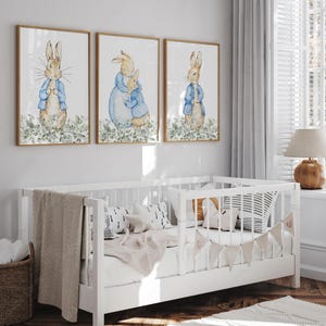 May include: Three framed watercolor prints featuring Peter Rabbit characters in blue jackets. The prints are displayed above a white crib with a beige blanket and decorative pillows. A woven basket and a lamp are also visible.