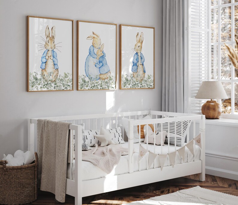 May include: Three framed watercolor prints featuring Peter Rabbit characters in blue jackets. The prints are displayed above a white crib with a beige blanket and decorative pillows. A woven basket and a lamp are also visible.