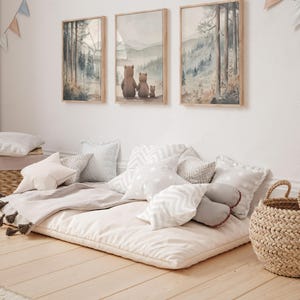 May include: Three framed watercolour prints of a forest scene and a family of bears hang above a floor cushion with pillows. The pillows are various shapes and patterns in shades of white and grey. A woven basket sits nearby.
