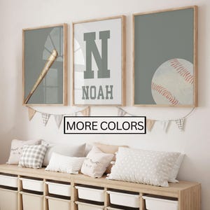 May include: Three framed prints with a baseball theme. One features a baseball bat, another a large letter "N" with the name "NOAH", and the third a baseball. The frames are light wood, and the background is a muted green. The text "MORE COLORS" is below the frames.