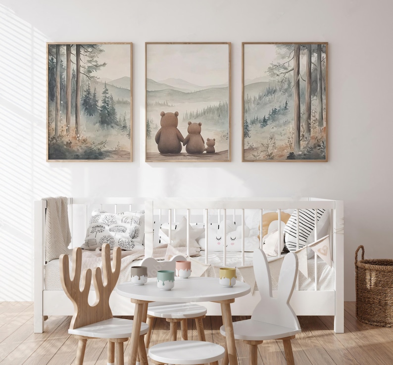 May include: Three framed watercolour prints showing a forest scene with a bear family, and a cot with decorative pillows. The prints have a soft colour palette of greens, browns, and blues. A small white table and chairs are in front of the cot.