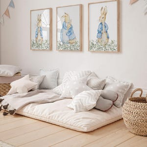 May include: Three framed watercolor prints of rabbits in blue jackets and a floor cushion with pillows. The prints are in wooden frames. The floor cushion is beige and surrounded by various pillows in white and gray.