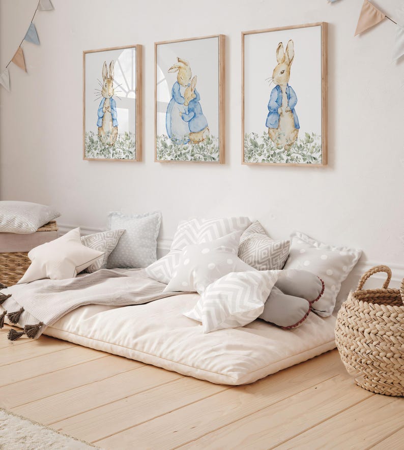 May include: Three framed watercolor prints of rabbits in blue jackets and a floor cushion with pillows. The prints are in wooden frames. The floor cushion is beige and surrounded by various pillows in white and gray.