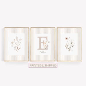 May include: Three framed floral art prints. The prints feature botanical illustrations in neutral tones, with a striped background. The center print displays the letter "E" with the name "Ellie" below. The frames have a gold finish.