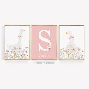May include: Three pink and white framed prints featuring a watercolour illustration of a white goose with a pink bow, a pink background with a white letter S and the name "Sophia", and a white goose with a pink bow. The prints are surrounded by a gold frame.