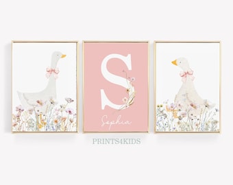 May include: Three pink and white framed prints with a watercolor illustration of a white goose with a pink bow, a pink background with a white letter S and the name "Sophia", and a white goose with a pink bow. The prints are surrounded by a gold frame.