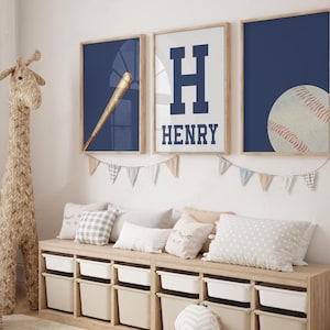 K&ouml;nnte beinhalten: Drei gerahmte Drucke mit einem marineblauen Hintergrund. Der erste Druck zeigt einen Baseballschl&auml;ger und einen Ball, der zweite Druck zeigt einen gro&szlig;en wei&szlig;en Buchstaben H mit dem Namen Henry darunter, und der dritte Druck zeigt einen Baseball. Die Drucke h&auml;ngen an einer wei&szlig;en Wand &uuml;ber einer Holzaufbewahrungsbank mit wei&szlig;en Beh&auml;ltern.