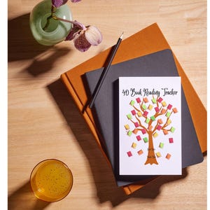 May include: A 40 Book Reading Tracker notebook with a tree design, featuring colorful book-shaped leaves. The notebook rests on top of a dark gray and orange notebook, with a pencil and a glass of orange liquid nearby. A vase with purple flowers is also present.