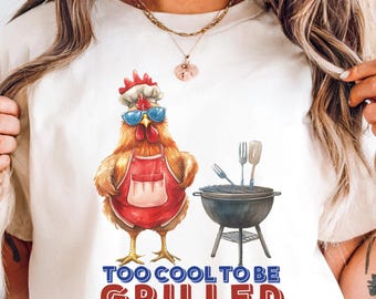 May include: Cream-colored t-shirt featuring a cartoon chicken wearing sunglasses, a chef's hat, and an apron, standing next to a grill. The text "TOO COOL TO BE GRILLED" is printed below the chicken in red and blue.