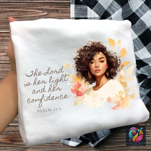 May include: White decorative pillow with a watercolor illustration of a woman with curly brown hair, gold hoop earrings, and floral accents. The text reads "The Lord is her light and her confidence" from Psalm 27:1.