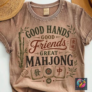 May include: A brown t-shirt with the words "GOOD HANDS, GOOD FRIENDS, GREAT MAHJONG" in a vintage style. The design includes bamboo stalks, decorative elements, and Mahjong tiles. The shirt has a crew neck and short sleeves.