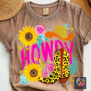 May include: Brown t-shirt with a colorful graphic. The design features the word "HOWDY" in pink, a yellow cowboy boot with a leopard print, a cowboy hat, sunflowers, and pink roses. The shirt has a vintage, faded look.