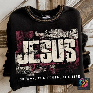 Op de afbeelding: Zwart sweatshirt met het woord "JESUS" in grote, versleten witte letters over een vervaagde bordeauxrode rechthoek. De tekst "THE WAY, THE TRUTH, THE LIFE" staat eronder. De kraag heeft een goudkleurige rand.