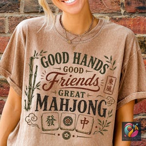 May include: A brown t-shirt with the text "GOOD HANDS GOOD FRIENDS GREAT MAHONG". The shirt features bamboo stalks, decorative flourishes, and Mahjong tiles. The shirt is a neutral color and has a vintage look.