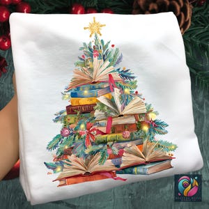 Könnte beinhalten: Weißes Sweatshirt mit einem Weihnachtsbaum-Design aus gestapelten Büchern, geschmückt mit festlichem Grün, Kugeln und einem goldenen Stern. Die Bücher sind geöffnet und zeigen ihre Seiten. Ein einzigartiges Feiertagsdesign.