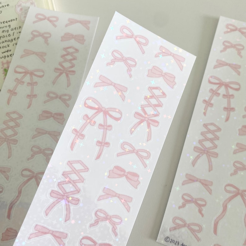 May include: A sheet of pink and white holographic stickers with various bow designs. The stickers have a glittery finish and are perfect for adding a touch of whimsy to planners, journals, and other craft projects.